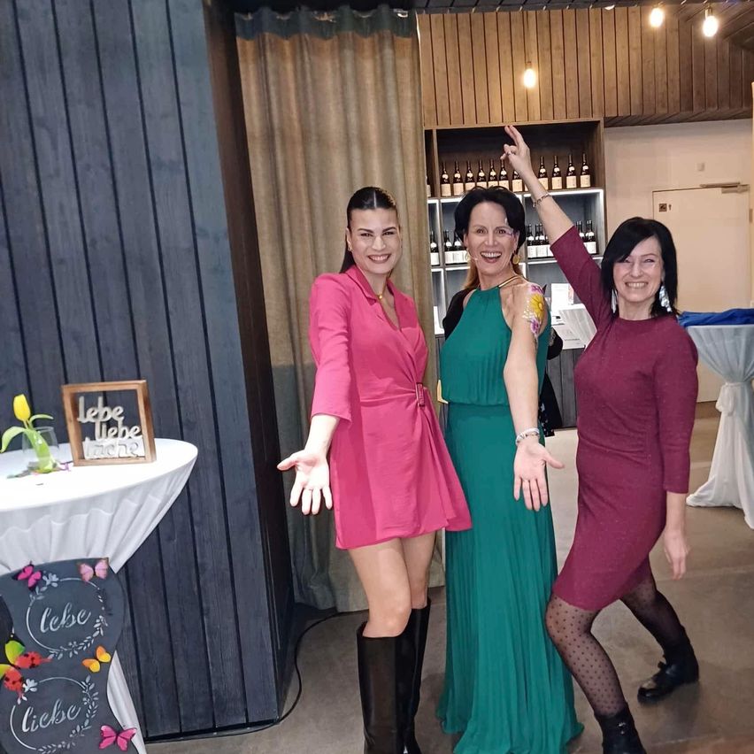 Drei Frauen stehen in einem Veranstaltungsraum zusammen. Sie tragen Kleider in Rosa, Grün und Lila. Eine Frau hat die Hand erhoben, und sie alle scheinen zu lächeln.