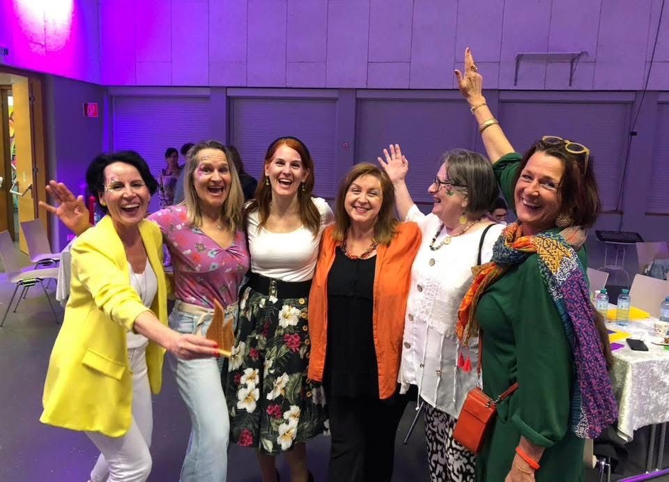 A group of women are smiling and posing for a photo with their hands up in a purple-lit room.