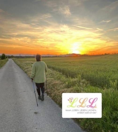 Eine ältere Frau geht bei Sonnenuntergang auf einem Landweg und stützt sich auf einen Stock. Der Himmel ist orange und pink gefärbt, während die Felder breit und grasig sind. Unten rechts ist ein Logo sichtbar.