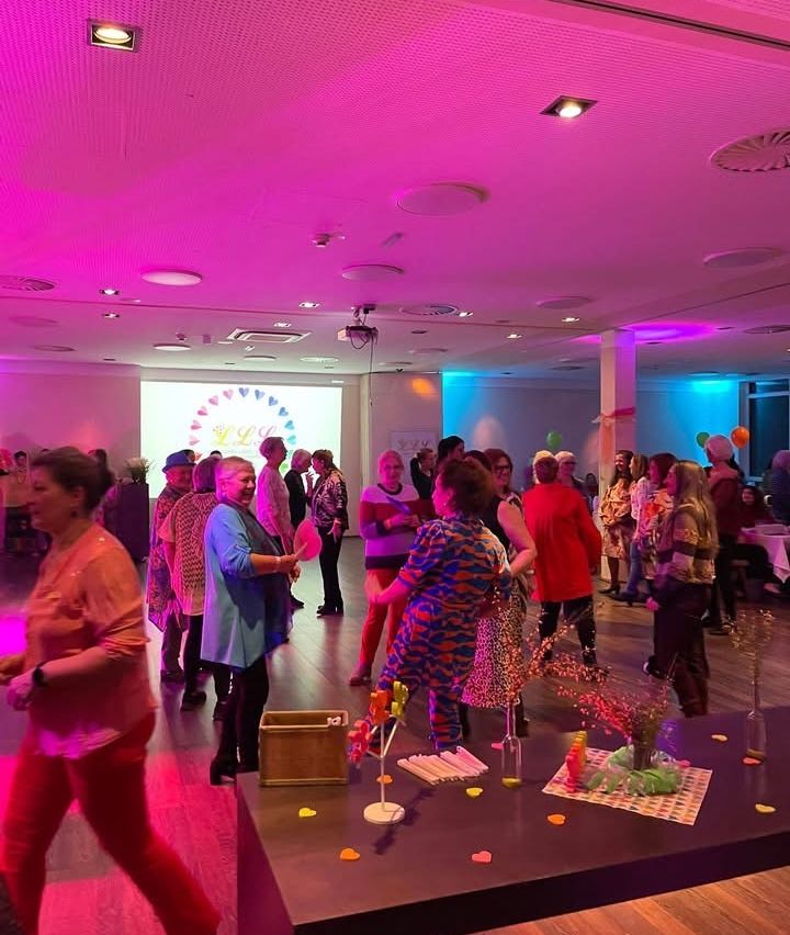 A group of people dressed in various outfits are standing and walking in a room with a projector screen and purple lighting. A table with flowers and decorations is in the foreground.