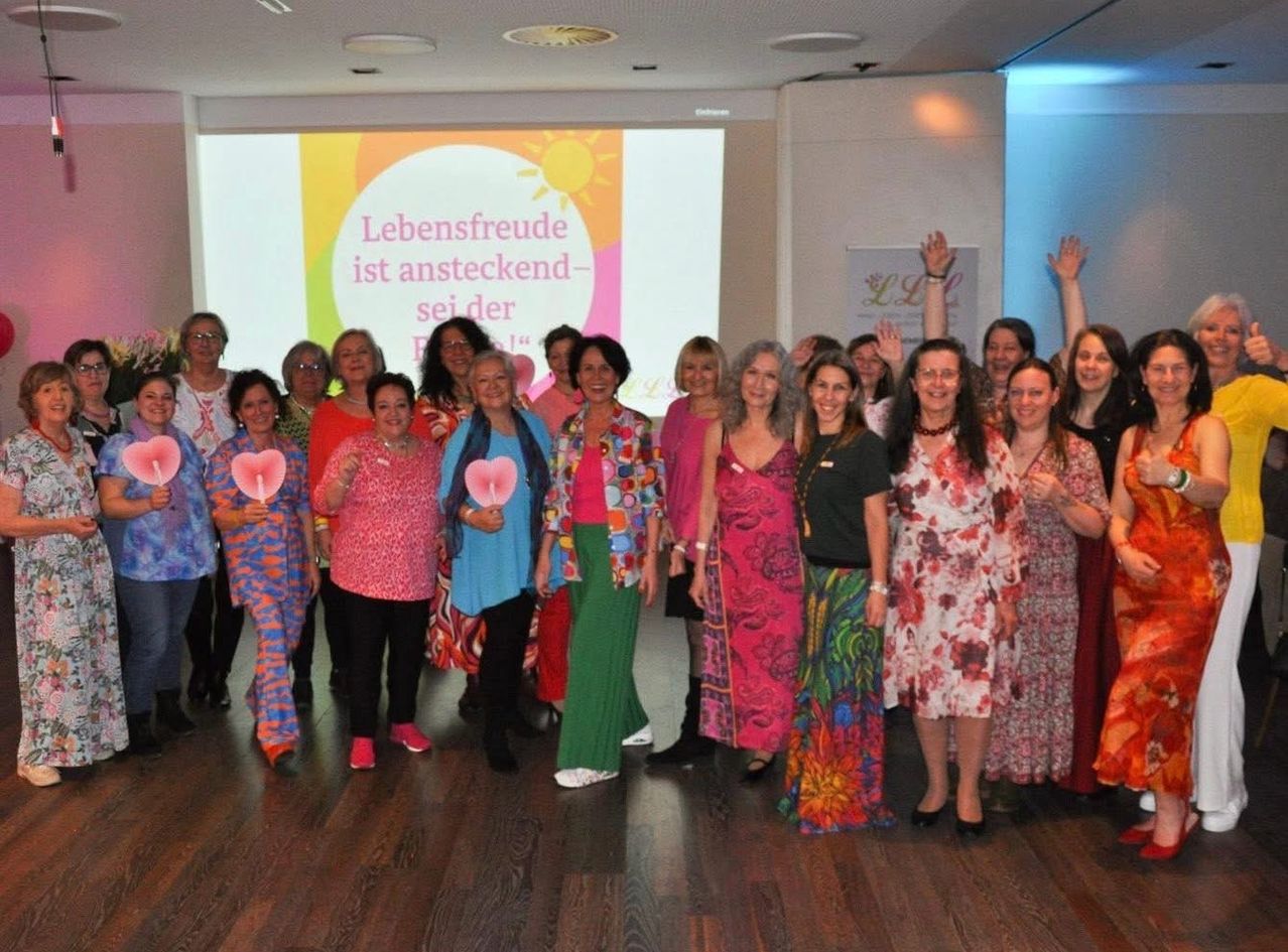 A group of women dressed in colorful attire poses for a photo in a room with a projector screen displaying a colorful message.
