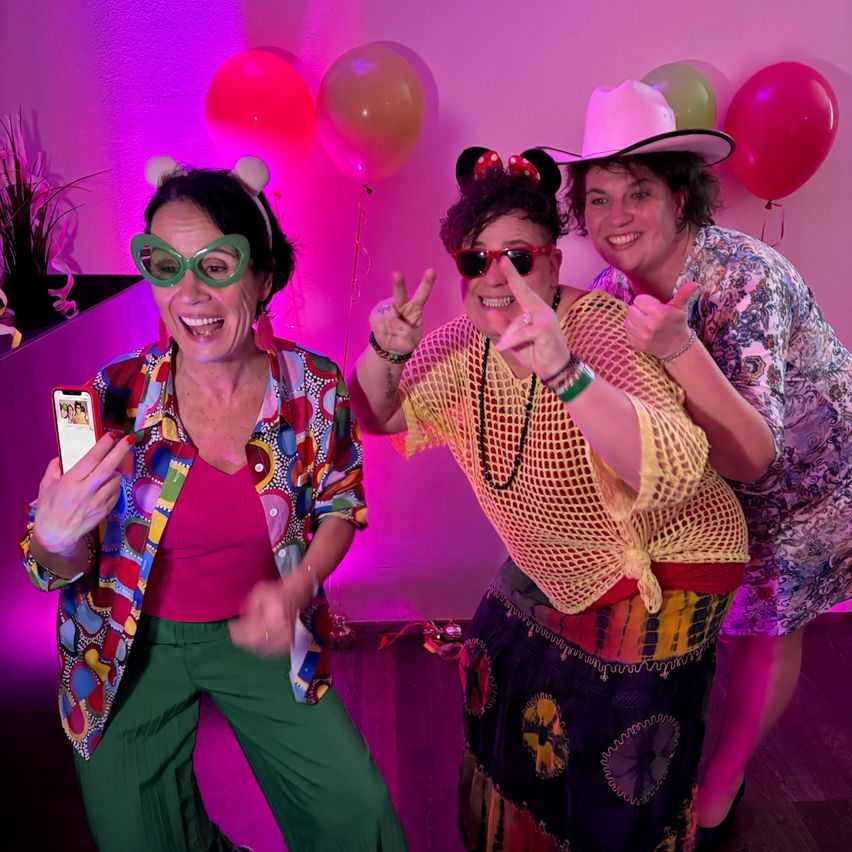 Three women pose with colorful attire and accessories, taking a selfie in a room with balloons and pink lighting.