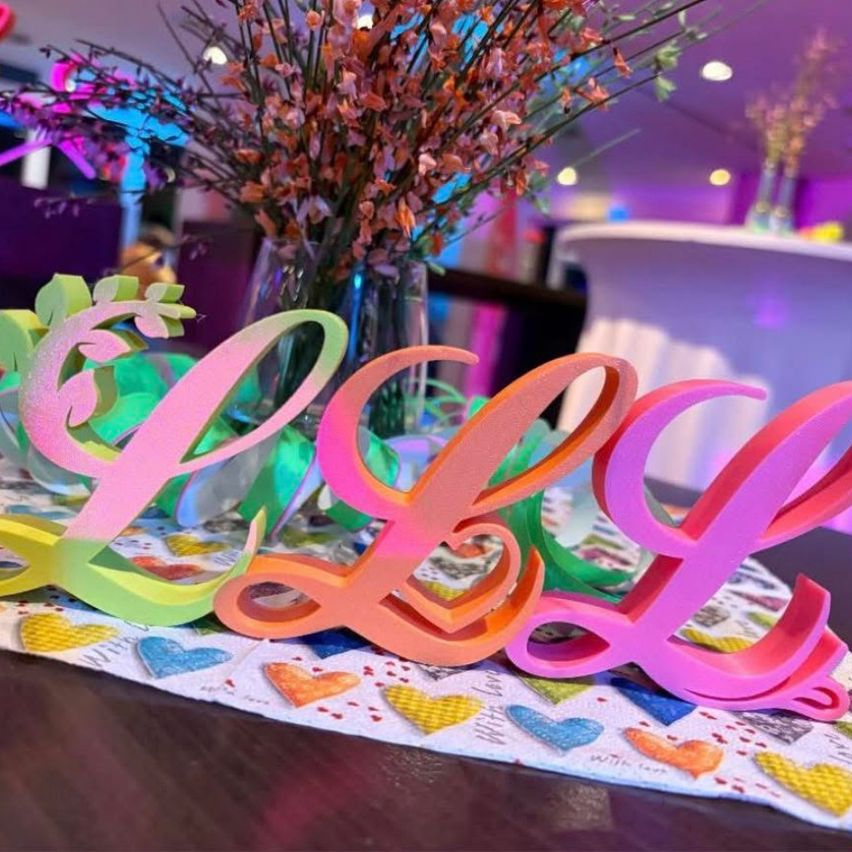 A table with a colorful floral centerpiece, a 'LLL' name sign in rainbow colors, and a heart-patterned tablecloth. The table is in a room with purple lighting.
