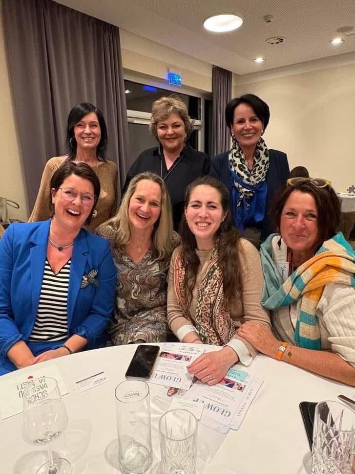 A group of women pose for a picture at a gathering. They sit around a table with pamphlets, smiling and looking at the camera. Behind them, a window and curtains are visible.