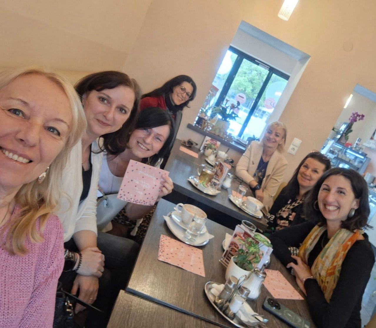 Six women seated around a table in a cafe, smiling for a photo. The table is set with cups, saucers, and a small potted plant. A window behind them shows a view of the outside.