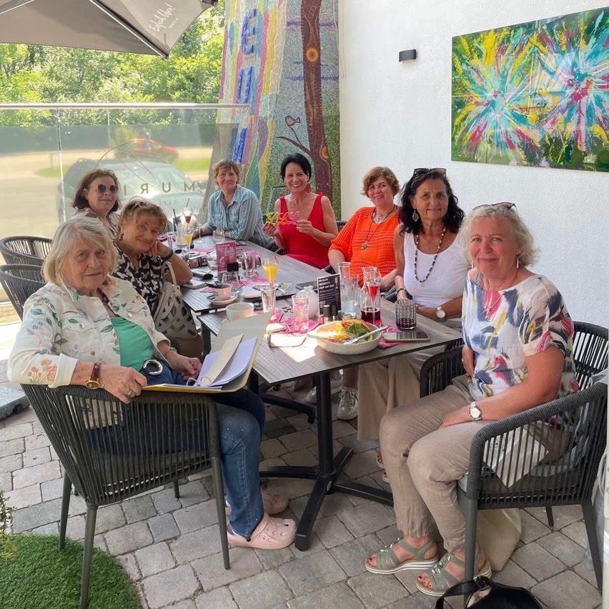 A group of women in casual attire are gathered around a table outside, possibly for a meeting. They are smiling and seem to be enjoying their drinks. The table has a bowl of food, glasses, and a menu. Behind them is a glass wall with a view of the outside.