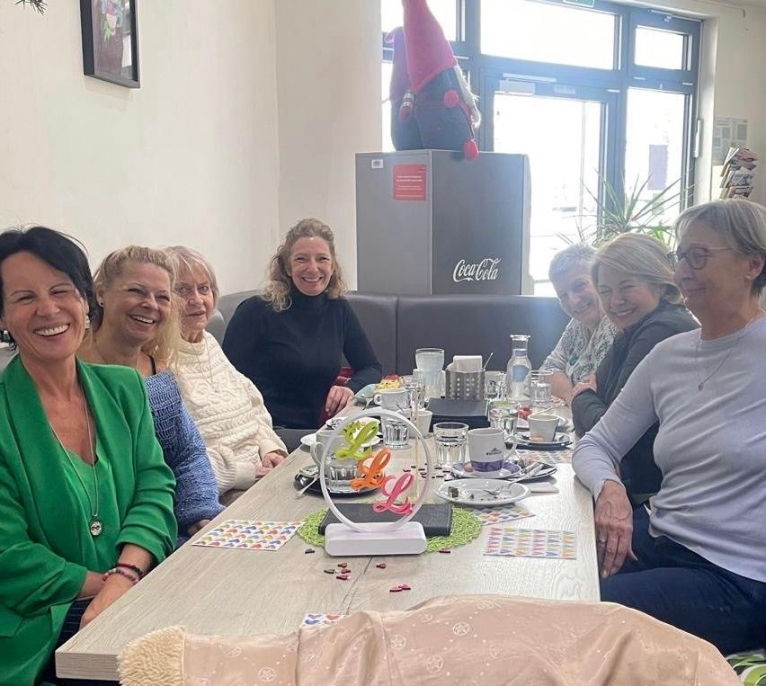 A group of women are seated around a table, smiling and posing for a photo. The table has cups, plates, and decorative items. Behind them is a refrigerator with a Coca-Cola logo and a gnome. A woman with glasses and a necklace sits at the end.
