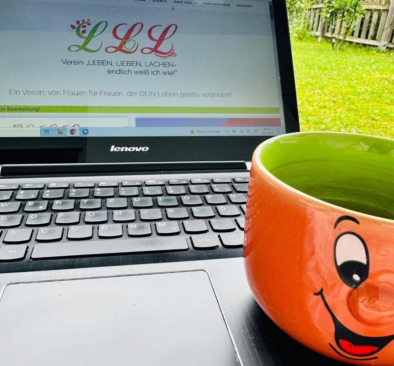 Ein Laptop zeigt eine Website mit einer lachenden Tasse daneben. Die Website zeigt 'LLL' mit einem farbenfrohen Logo und einem Motto. Die Tastatur ist im Vordergrund.