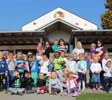 Eine Gruppe von Kindern und Erwachsenen steht vor einem Gebäude mit einem Regenbogendesign auf dem Dach. Einige von ihnen tragen Brillen und einige tragen Babys. Sie posieren für ein Foto.