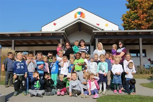 Eine Gruppe von Kindern und Erwachsenen steht vor einem Gebäude mit einem Regenbogendesign auf dem Dach. Einige von ihnen tragen Brillen und einige tragen Babys. Sie posieren für ein Foto.