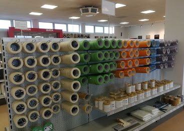 Ein Geschäft zeigt eine Vielzahl farbenfroher Klebebänder, die an einer Lochwand montiert sind. Regale halten zusätzliche Bänder und Dosen. Die Decke ist mit weißen Leuchten ausgestattet.