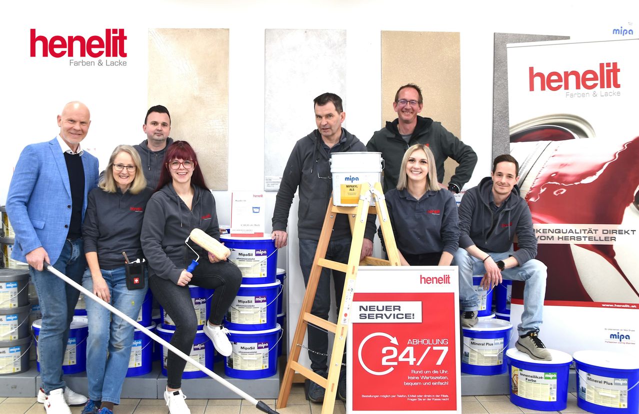 Eine Gruppe von Menschen steht vor einer Wand mit Mustertafeln. Sie halten Eimer, Pinsel und Farbwalzen. Eine Frau hält ein rotes Schild mit der Aufschrift '24/7 Service'. Ein Mann hält einen weißen Eimer mit dem Wort 'Mipa' darauf.