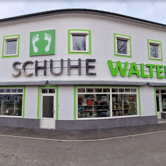 Die Vorderseite eines weißen Gebäudes mit grünen Akzenten, beschriftet mit 'Schuhe Walter', hat mehrere Fenster und eine Tür. Im Inneren sind Schuhe ausgestellt.