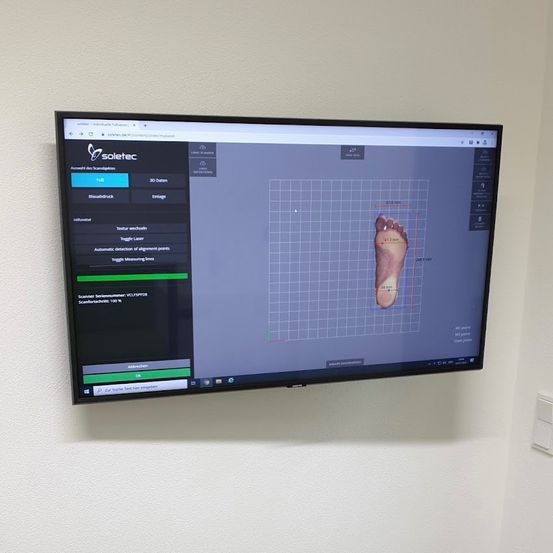 Ein Monitor zeigt eine Fußscan-Oberfläche mit 'soletec'-Branding und Optionen wie 'Scan', '3D-Daten', 'Erstellen', 'Messung' und 'Einstellungen'. Der Scan zeigt eine Fußkontur mit Messlinien und Datenpunkten.