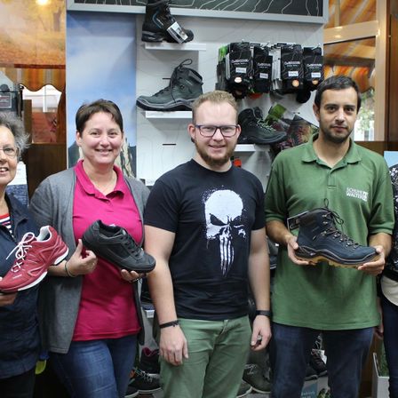 Fünf Menschen posieren für ein Foto in einem Schuhgeschäft und halten verschiedene Schuhe in der Hand. Drei Männer und zwei Frauen stehen eng beieinander und lächeln, mit Regalen voller Stiefel und Socken im Hintergrund.