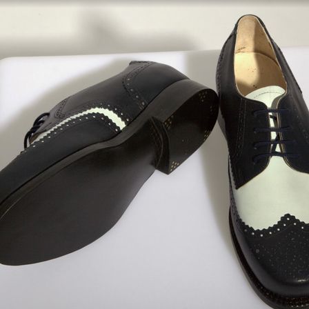 Zwei schwarze Lederschuhe mit weißen Besätzen und dekorativen Löchern liegen nebeneinander auf einer weißen Oberfläche. Der linke Schuh hat eine schwarze Sohle und ein weißes Inneres. Der rechte Schuh hat eine weiße Sohle und ein schwarzes Inneres.