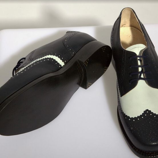 Zwei schwarze Lederschuhe mit weißen Besätzen und dekorativen Löchern liegen nebeneinander auf einer weißen Oberfläche. Der linke Schuh hat eine schwarze Sohle und ein weißes Inneres. Der rechte Schuh hat eine weiße Sohle und ein schwarzes Inneres.