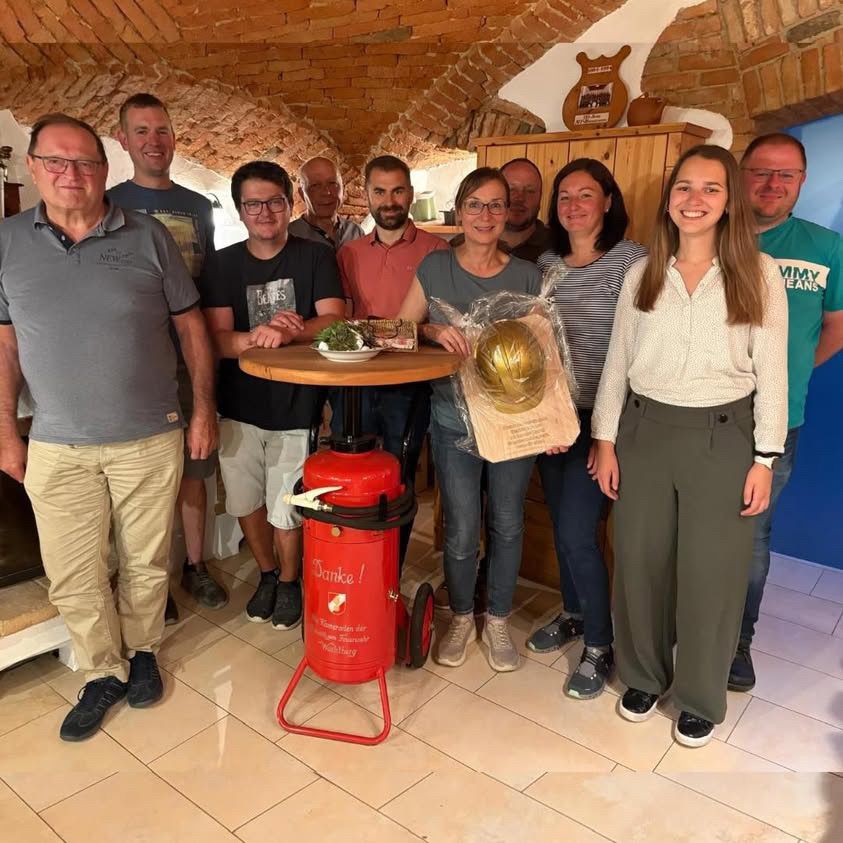 Eine Gruppe von Menschen steht in einem Raum mit Ziegelwänden, lächelt und posiert für ein Foto. Eine Frau hält eine Trophäe und ein Mann einen roten Feuerlöscher. In der Mitte steht ein Tisch mit einer Pflanze.