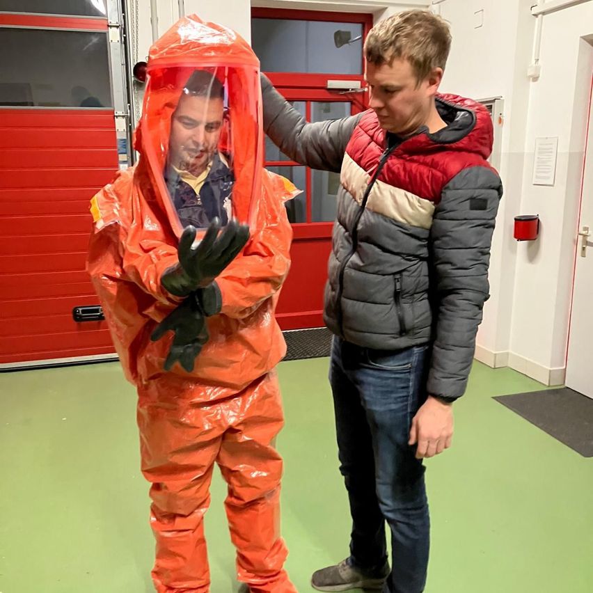 Ein Mann in einem roten Schutzanzug und ein Mann in einer Jacke stehen in einem Raum mit grünem Boden. Der Mann im Schutzanzug lächelt.