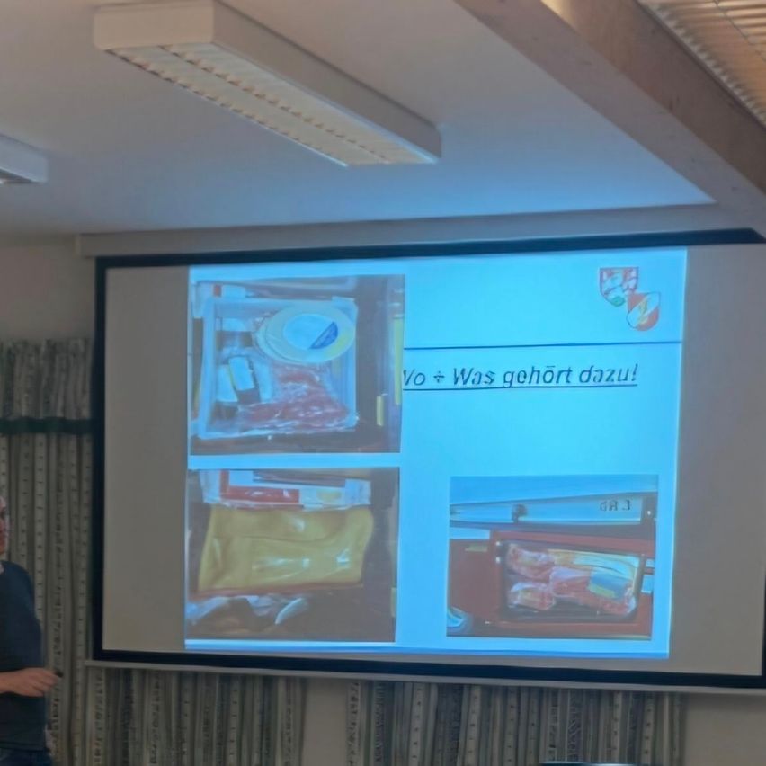 Eine Projektionsleinwand zeigt eine Folie mit den Worten 'Yo + Was gehort dazu!' und Bildern von Lebensmitteln in Plastikbehältern. Eine Person steht neben der Leinwand.