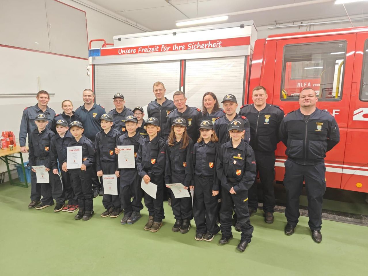 Eine Gruppe von Feuerwehrleuten, sowohl Männer als auch Frauen, steht hinter einem Feuerwehrauto. Sie posieren für ein Foto mit Kindern, die Urkunden halten. Das Feuerwehrauto trägt die Aufschrift 'Unsere Freiheit für Ihre Sicherheit'.