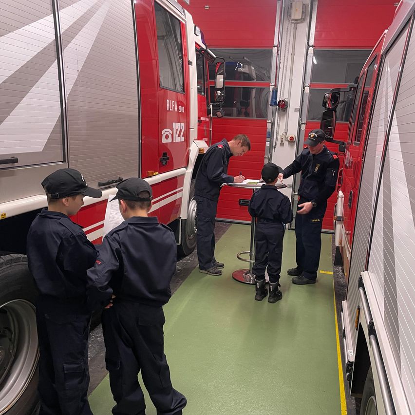Eine Gruppe junger Jungen in Feuerwehruniformen beobachtet einen Mann, der ein Dokument auf einem kleinen Tisch in einer Feuerwache unterzeichnet. Zwei Feuerwehrleute stehen hinter dem Tisch, einer trägt einen Hut.