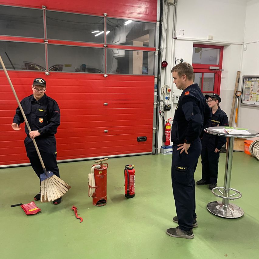 Drei Männer in Uniformen stehen in einer Garage. Einer hält eine Kehrschaufel, ein anderer beobachtet ihn, und ein Dritter steht hinter einem Tisch. Feuerlöscher und eine Feuerhydrante sind in der Nähe.