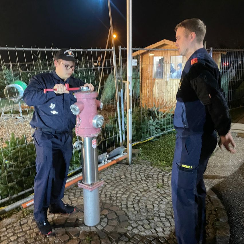 Zwei Männer in Marineblau-Uniformen arbeiten bei Nacht an einem Hydranten in der Nähe eines Zauns. Hinter ihnen befindet sich ein kleines Holzhaus mit einem Fenster.