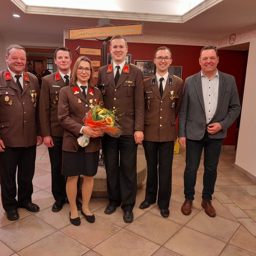 Fünf Personen in Uniform stehen zusammen in einem Raum. Eine Frau in der Mitte hält einen Blumenstrauß. Hinter ihnen steht ein Schild mit der Aufschrift 'Gasthof am Bahnhof.'
