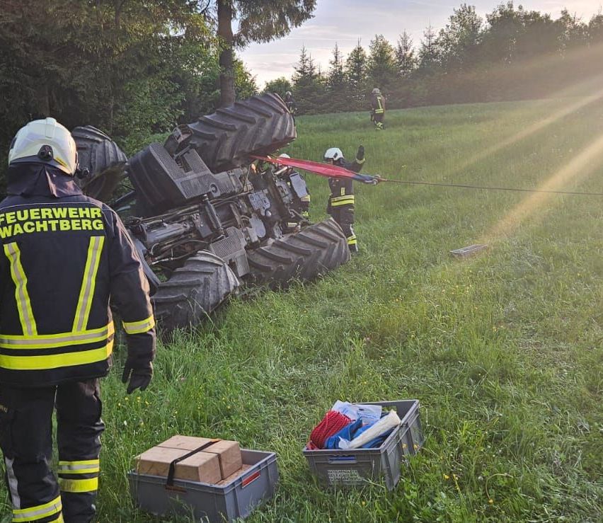 Feuerwehrleute ziehen ein großes Fahrzeug aus einem Grasfeld. Ein Feuerwehrmann hält ein Seil, während zwei andere ziehen. Lagerboxen sind neben ihnen.