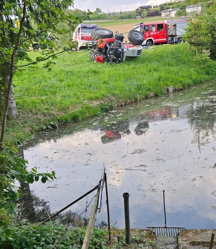 Ein Traktor ist in einem Feld neben einem Bach umgekippt. Feuerwehrwagen sind in der Nähe geparkt, und eine Person steht neben dem Traktor.