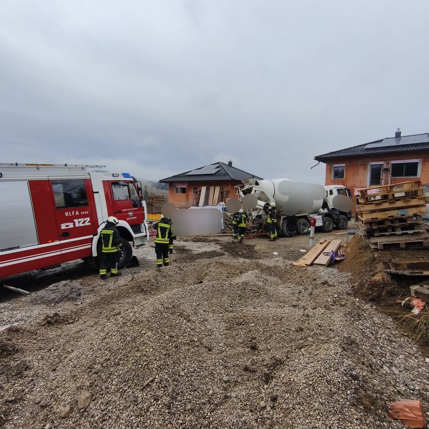 Feuerwehrleute stehen auf einer Baustelle mit einem großen Zementlastwagen und einer weißen Struktur. Zwei Häuser sind im Hintergrund.