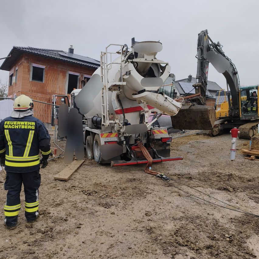 Ein Feuerwehrmann in voller Montur steht in der Nähe einer Baustelle. Ein Zementmischer ist festgefahren, und ein Bagger ist in der Nähe. Hinter dem Zaun steht ein Haus im Bau.