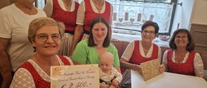 Eine Gruppe von Frauen in traditioneller Kleidung, die lächelnd für ein Foto posieren. Eine Frau hält ein Baby, eine andere eine Urkunde mit einem Geldbetrag. Hinter ihnen ein Fenster mit Blick auf ein Auto und Bäume.