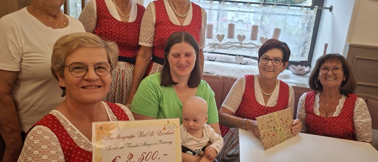 Eine Gruppe von Frauen in traditioneller Kleidung, die lächelnd für ein Foto posieren. Eine Frau hält ein Baby, eine andere eine Urkunde mit einem Geldbetrag. Hinter ihnen ein Fenster mit Blick auf ein Auto und Bäume.