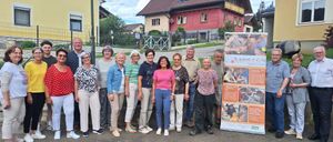 Eine Gruppe von Erwachsenen, sowohl Männer als auch Frauen, steht vor Häusern, einige mit Brille, alle lächeln für ein Foto. Ein Schild ist hinter ihnen.