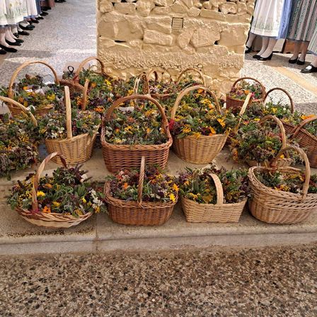 Eine Reihe von Weidenkörben gefüllt mit getrockneten Blumen steht auf einer Steinfläche vor einer Steinmauer. Im Hintergrund stehen Menschen in traditioneller Kleidung.