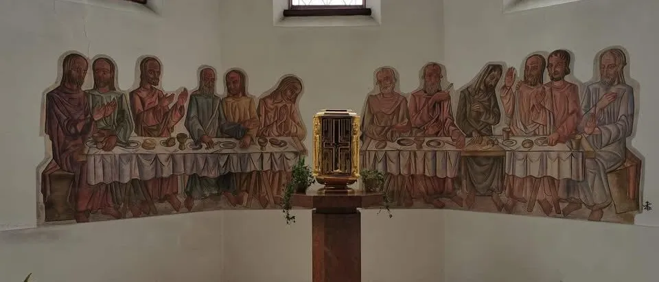 Eine Kapelle mit einem Letztes-Abendmahl-Wandbild und einer goldenen Box auf einem Holzständer. Die Wände sind weiß gestrichen, und es gibt zwei Fenster mit braunen Rahmen.