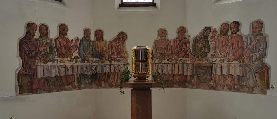 Eine Kapelle mit einem Letztes-Abendmahl-Wandbild und einer goldenen Box auf einem Holzständer. Die Wände sind weiß gestrichen, und es gibt zwei Fenster mit braunen Rahmen.