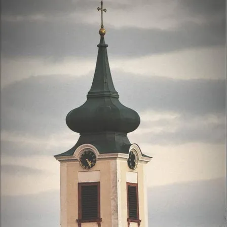 Ein Uhrturm erhebt sich gegen einen bewölkten Himmel. Er hat eine grüne Kuppel, zwei Uhren und einen Turm. Darunter ein Gebäude mit einem braunen Dach und einem Baum davor.