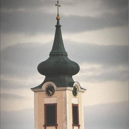 Ein Uhrturm erhebt sich gegen einen bewölkten Himmel. Er hat eine grüne Kuppel, zwei Uhren und einen Turm. Darunter ein Gebäude mit einem braunen Dach und einem Baum davor.
