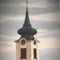 Ein Uhrturm erhebt sich gegen einen bewölkten Himmel. Er hat eine grüne Kuppel, zwei Uhren und einen Turm. Darunter ein Gebäude mit einem braunen Dach und einem Baum davor.