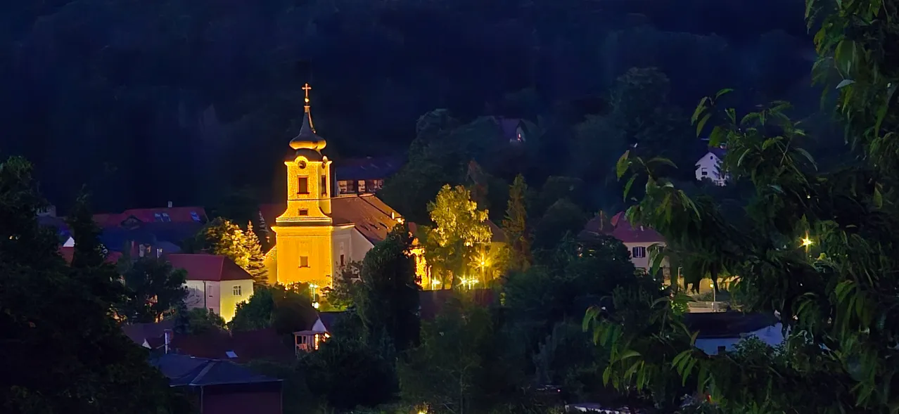 Eine gelbe Kirche mit einem Turm ist nachts beleuchtet, umgeben von Bäumen und Häusern. Die Kirche ist mit warmem Licht beleuchtet und es gibt mehrere Fenster.