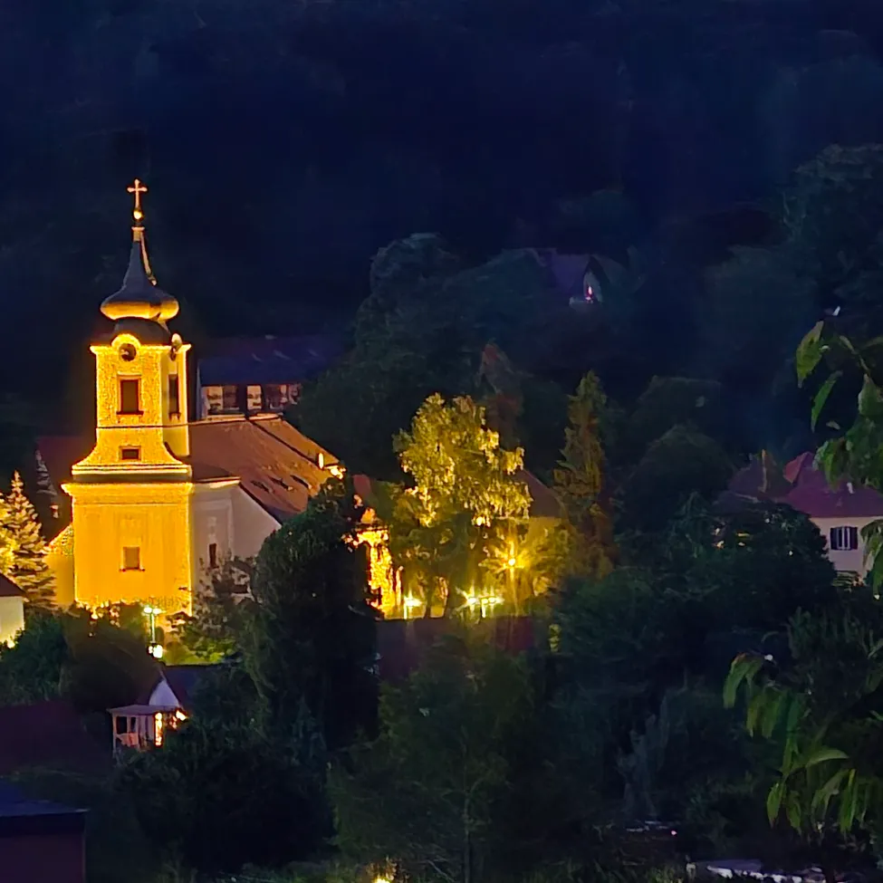 Eine gelbe Kirche mit einem Turm ist nachts beleuchtet, umgeben von Bäumen und Häusern. Die Kirche ist mit warmem Licht beleuchtet und es gibt mehrere Fenster.