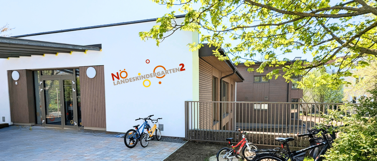 Ein weisses Gebäude mit Landeskindergarten 2-Schild, vor dem Fahrräder geparkt sind, und einem Metallzaun. Bäume und Gras umgeben das Gebiet.