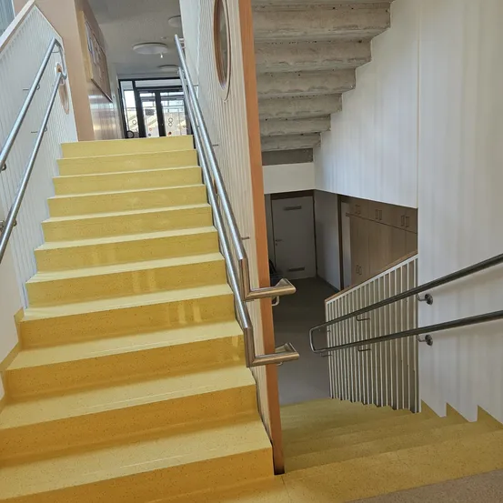 Gelbe Treppen mit Metallhandläufen in einem modernen Gebäude. Die Treppe führt zu einer Tür im oberen Stockwerk.