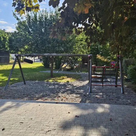 Ein Spielplatz im Freien mit Schaukeln, einer Rutsche und Klettergerüst. Bäume und Gras umgeben das Gebiet.