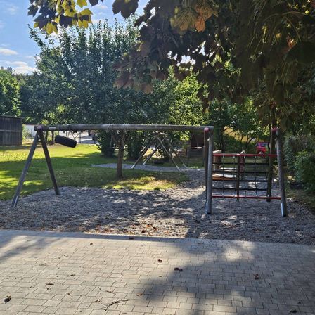 Ein Spielplatz im Freien mit Schaukeln, einer Rutsche und Klettergerüst. Bäume und Gras umgeben das Gebiet.