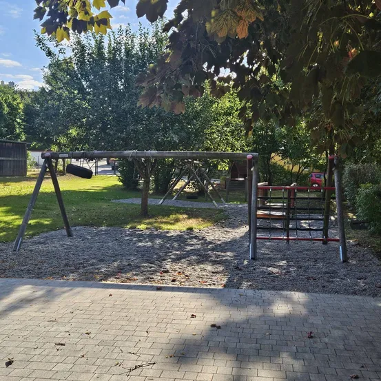 Ein Spielplatz im Freien mit Schaukeln, einer Rutsche und Klettergerüst. Bäume und Gras umgeben das Gebiet.