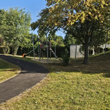 Ein Außenspielplatz mit Rutsche, Klettergerüst und einem gepflasterten Weg. Der Bereich ist von Rasen und Bäumen umgeben. Ein weißes Gebäude ist im Hintergrund zu sehen.